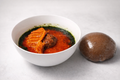 Bowl of soup with a piece of meat and a dark brown ball on a light gray background