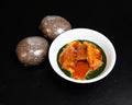 A white bowl containing a green and orange-red food dish with pieces on top, placed on a black background next to two brown round items with white powder dusting.