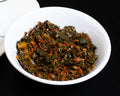  a small white bowl filled with a dark green and red seasoned vegetable mixture, set against a solid black background.