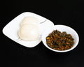 Two steamed white buns on a white square plate next to a small white bowl filled with a dark green and red seasoned vegetable mixture, set against a solid black background.