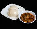 Two steamed buns on a white square plate next to a small white bowl of spicy vegetable sauce, set against a solid black background.