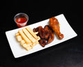 A white rectangular plate featuring golden french fries, grilled chicken wings, and a glazed chicken drumstick, served with a small clear plastic cup of red dipping sauce on a black background.