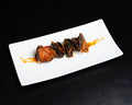 A white rectangular ceramic plate holding several dark, reddish-brown food pieces arranged in a row with orange sauce drizzle, photographed on a black background.