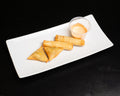 Four golden-brown spring rolls and two triangular samosas served on a white rectangular plate with a small clear cup of orange dipping sauce, set against a solid black background.