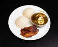 A white plate of food on a black background, featuring two balls of white rice, two strips of fried plantain, and a small clear plastic cup of green curry with a halved boiled egg.